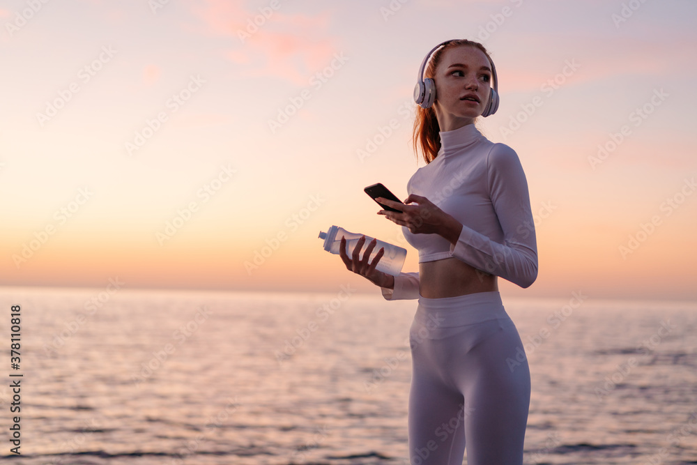 Image of young redhead sportswoman in headphones using mobile phone