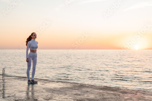 Image of redhead focused sportswoman in earphones standing on promenade
