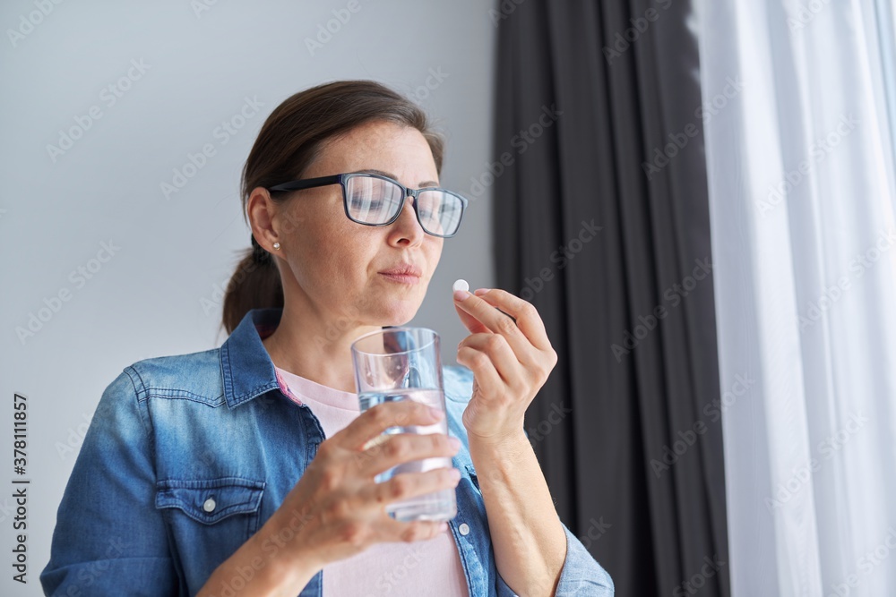 Mature middle-aged woman at home holding pill and glass of fresh water Mature middle-aged woman at home holding pill and glass of fresh water