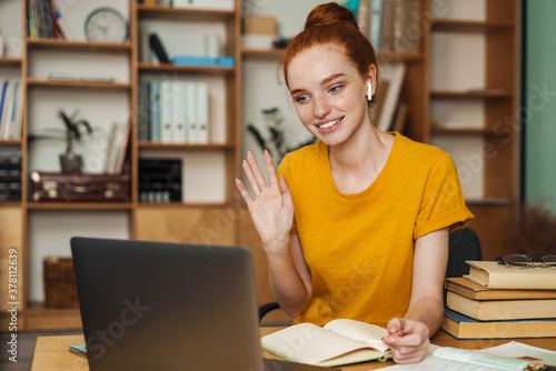 Image of smiling girl waving hand while doing homework with laptop