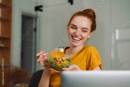 Image of laughing redhead girl eating salad while working with laptop