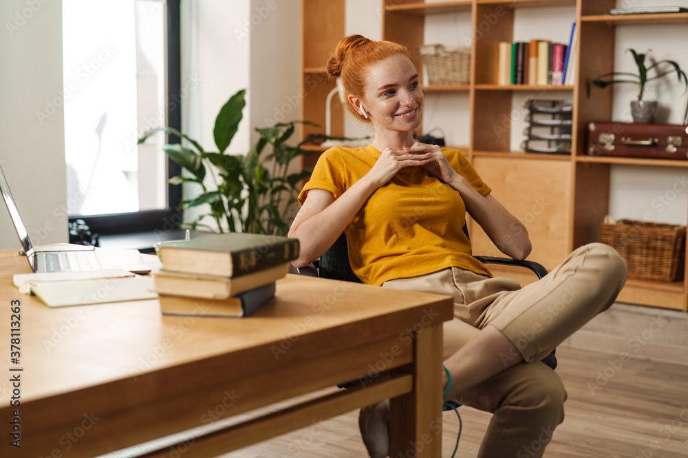 Image of smiling redhead girl using earphones while working with laptop Image of smiling redhead girl using earphones while working with laptop