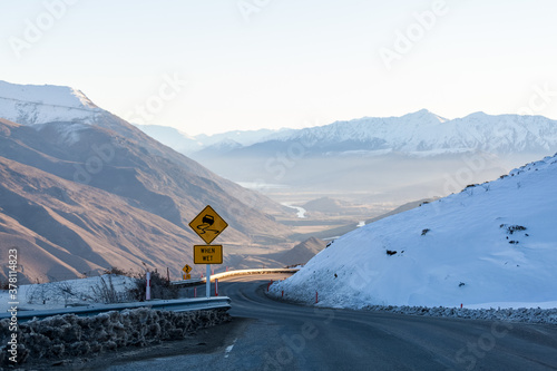 Fototapeta Naklejka Na Ścianę i Meble -  A winding road leads down the snow covered Crown Ranges, a slippery when wet road sign on the left warning drivers to take care during their travel. Arrow Junction, New Zealand.