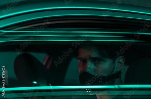 a young man in a face mask driving a car in the night city with neon lights reflecting from the glass of his car