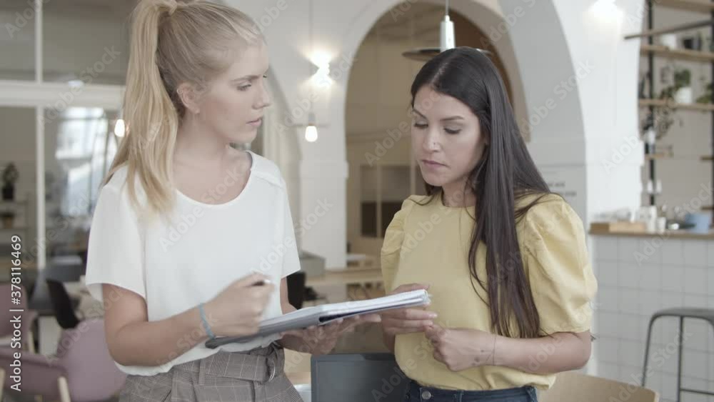 Serious female project colleagues discussing document. Young women pointing at papers and talking while standing in co-working space. Medium shot. Teamwork or paperwork concept Serious female project colleagues discussing document. Young women pointing at papers and talking while standing in co-working space. Medium shot. Teamwork or paperwork concept