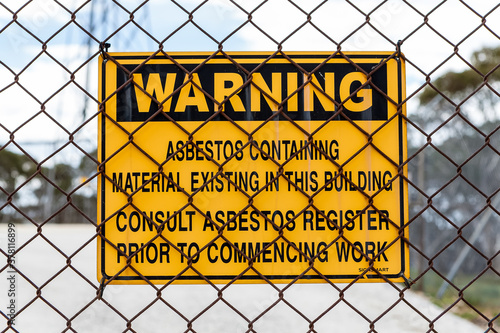 An asbestos warning sign attached to a rusty wire fence alerting those entering to the possible danger in the nearby buildings.