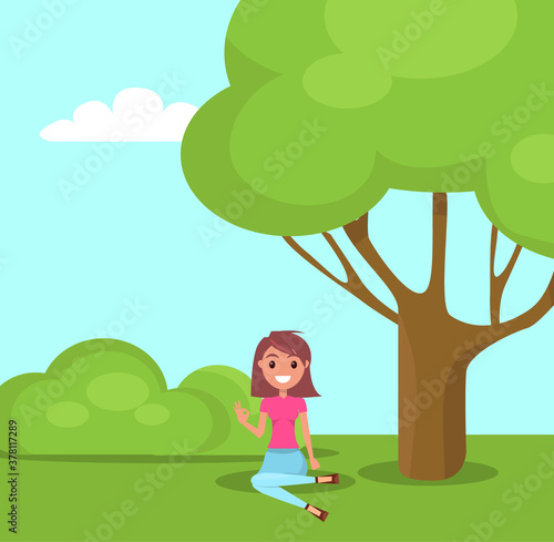 Young girl sitting at green grass in park near tree, smiling stylish girl relaxing at nature, summer time. Pretty female wearing t-shirt, jeans have recreation. Woman with bob hairstyle show ok sign