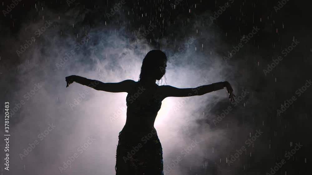 Dark silhouette of exotic dancer on a smoky background in a dark studio. A woman dances seductively among the raindrops in an oriental dress. Slow motion. Close up. Dark silhouette of exotic dancer on a smoky background in a dark studio. A woman dances seductively among the raindrops in an oriental dress. Slow motion. Close up.