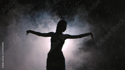 Dark silhouette of exotic dancer on a smoky background in a dark studio. A woman dances seductively among the raindrops in an oriental dress. Slow motion. Close up.