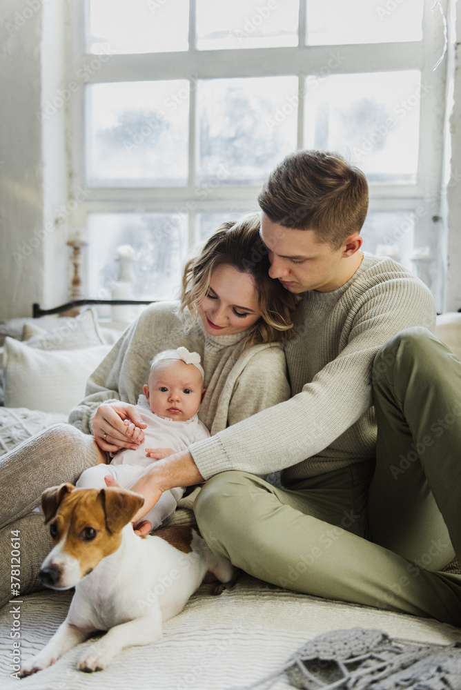 Young women and a man hold a newborn in their arms in the room near the Christmas tree. Family with a newborn and a dog on the bed in a Christmas interior. Young women and a man hold a newborn in their arms in the room near the Christmas tree. Family with a newborn and a dog on the bed in a Christmas interior.