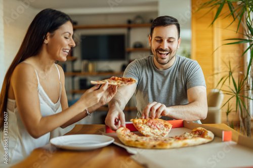 Positive couple eating pizza while sitting at kitchen table at home.