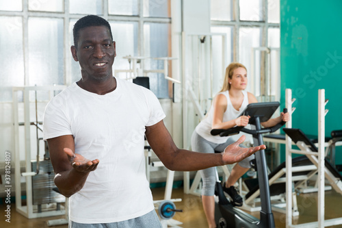 Portrait of positive friendly afro man welcoming to gym with exercising woman
