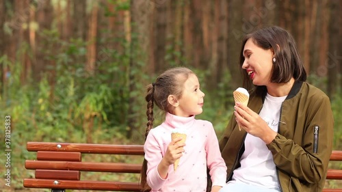Cute toddler girl and mother eating ice-cream sitting on bench in the park