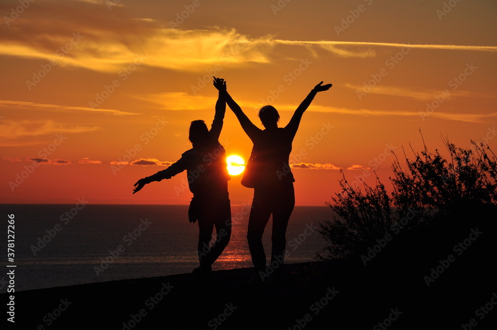 Silhouettes of two girls at sunset who stand on a mountain overlooking the sea. Vacation  concept.