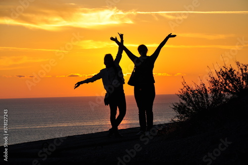 Silhouettes of two girls at sunset who stand on a mountain overlooking the sea. Vacation and tourism concept.