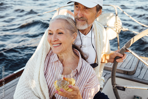 Elderly couple sitting together on a private yacht and wrapped in a plaid. Smiling caucasian woman drinking a wine while her husband stearing a wheel. © Artem Varnitsin