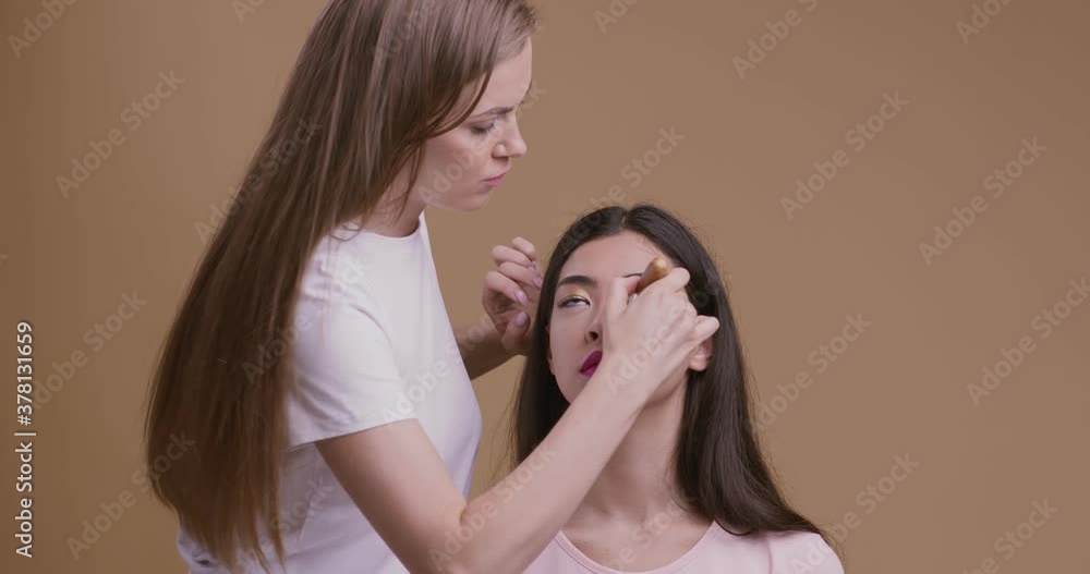 Make up artist finishing glam look for young asian woman at salon, beige studio background Make up artist finishing glam look for young asian woman at salon, beige studio background