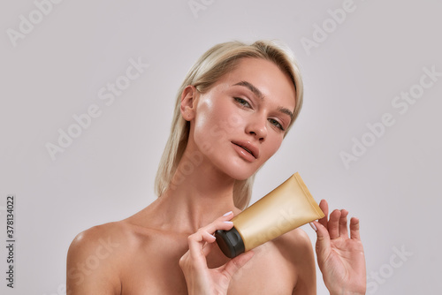 Portrait of young woman with perfect glowing skin looking at camera  holding sunscreen skincare product while posing isolated over grey background
