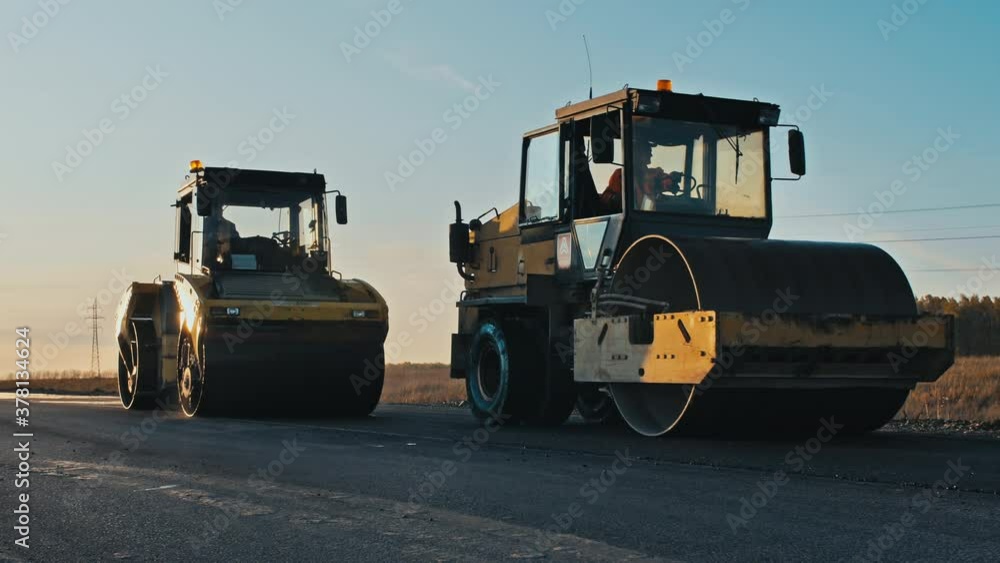 Road surface repair. Construction of a new road. Three rollers level and compact the asphalt at sunset, cars pass by. A layer of freshly laid asphalt in the rays of the setting sun. Road surface repair. Construction of a new road. Three rollers level and compact the asphalt at sunset, cars pass by. A layer of freshly laid asphalt in the rays of the setting sun.