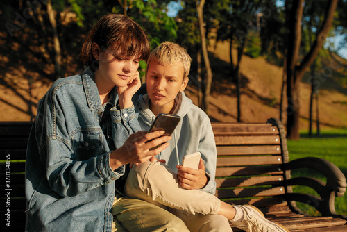 Two young girls holding their smartphone, sitting on the bench in the city park. Lesbian couple using their phones while spending time together outdoors © Svitlana