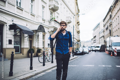 Serious young hipster male talking on mobile phone while walking on city road
