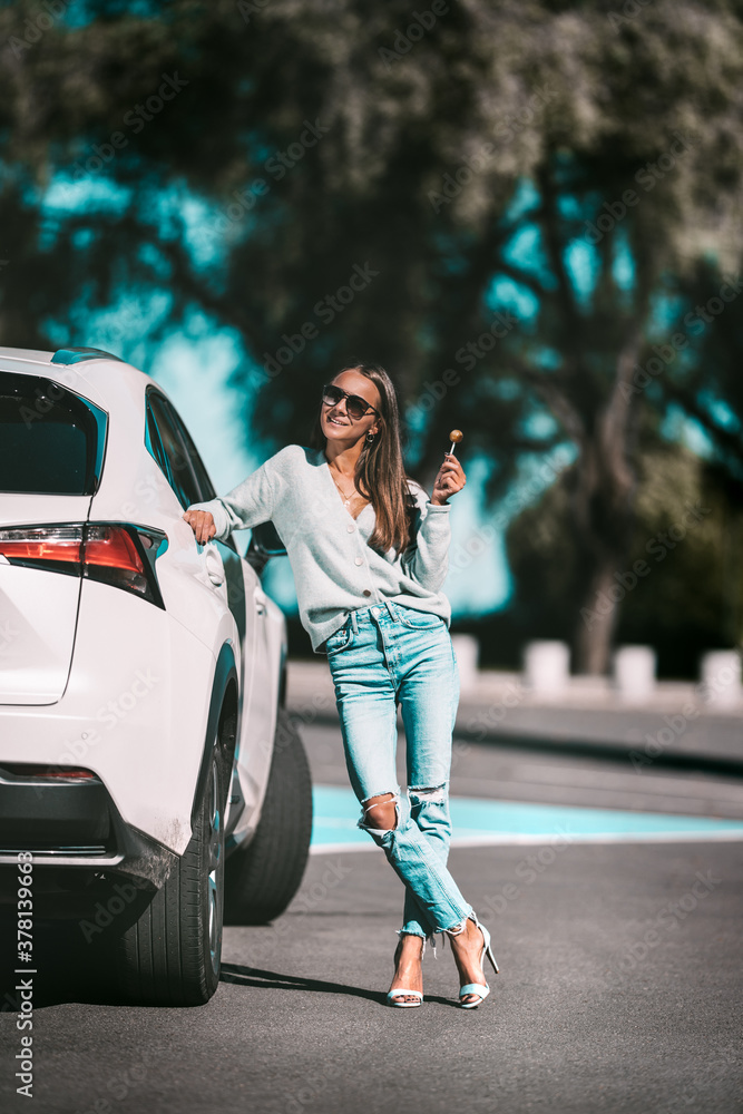 Adorable fit brunette woman in stylish clothes with lollipop stands near her new white electro car in sunny summer day. Successful young businesswoman. Adorable fit brunette woman in stylish clothes with lollipop stands near her new white electro car in sunny summer day. Successful young businesswoman.