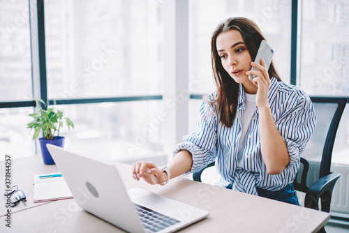 Smart casual female employee using smartphone app for making service consultancy during laptop programming in modern workspace, Caucasian woman connecting to 4g for phoning and call talking via mobile