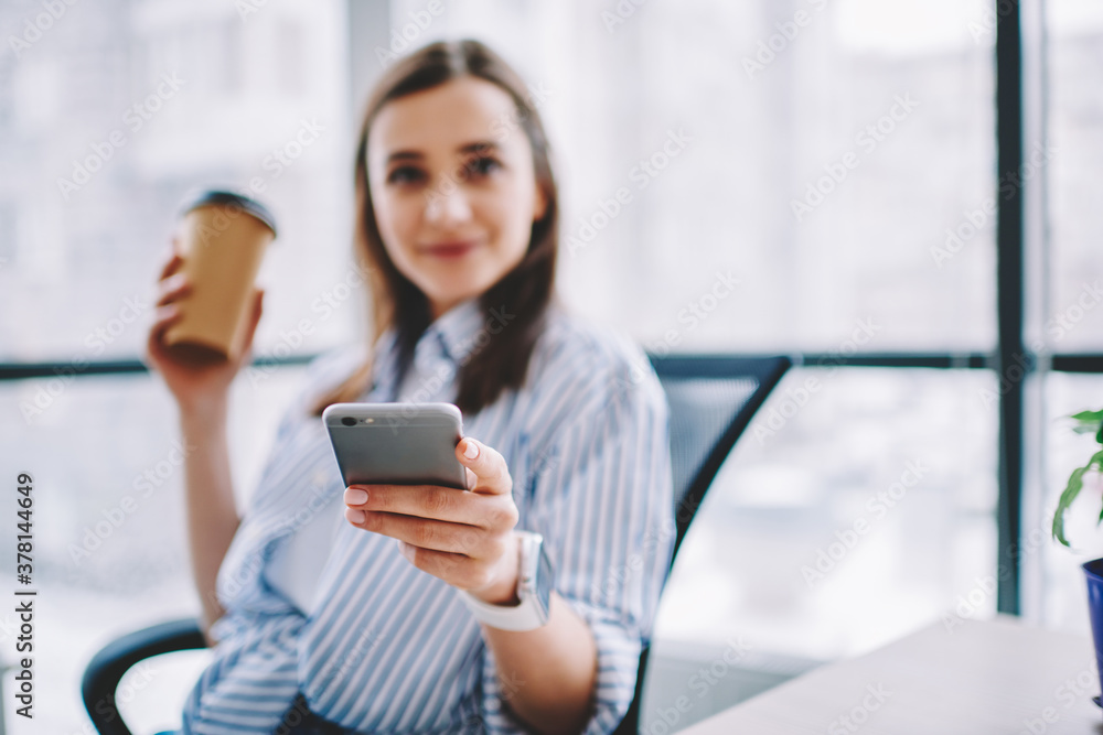 Portrait of blurred millennial using cellular technology during coffee break in office, selective focus on female hand holding modern smartphone device for wireless browsing via application
