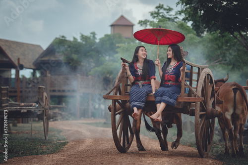 The way of life of Thai women from ancient times in traditional Thai costumes, sitting on a bullock cart or riding a cart. The backdrop is a beautiful ancient wooden house in Thailand. © เลิศลักษณ์ ทิพชัย
