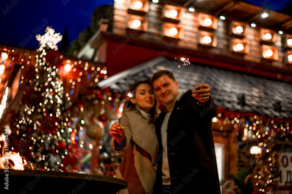 Couple in love relationship believe in meracle, kissing against the chrismas decoration outdoor in the eve, look at each other and holding sparklers Couple in love relationship believe in meracle, kissing against the chrismas decoration outdoor in the eve, look at each other and holding sparklers