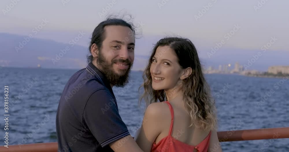 Young lovers are watching sea and turning to camera, smiling. Happy couple concept, togetherness, happiness and love. Young lovers are watching sea and turning to camera, smiling. Happy couple concept, togetherness, happiness and love.