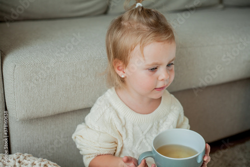 A little cute girl in an orange sweater is drinking tea. Cozy portrait of a girlsitting at home. Fall. photo