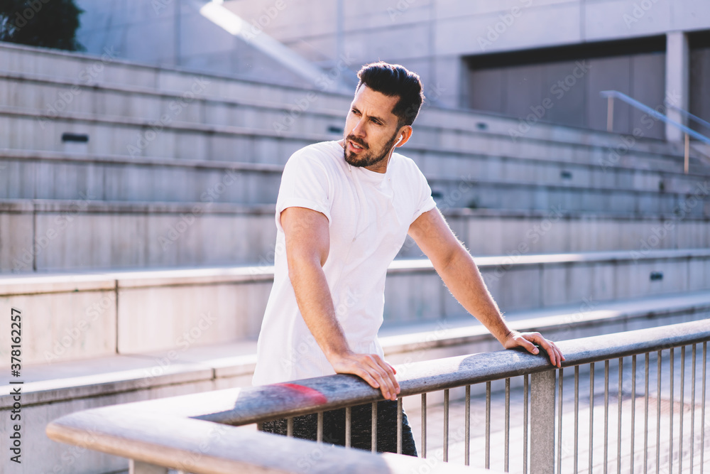 Thoughtful Caucasian male in electronic headphones listening sportive audio podcast during workout break at urban setting, pensive man 30s pondering on physical training goals for practice