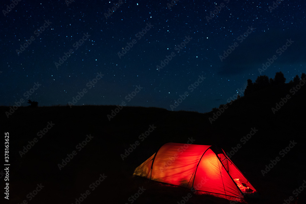 Glowing red tent with hikers sleeping in the wild mountains under dramatic night sky. Lit camping tent in the forest under starry sky during summer vacation.