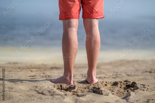Barefoot male feet stand on a sandy beach, against a blurred ocean background, on a sunny day.