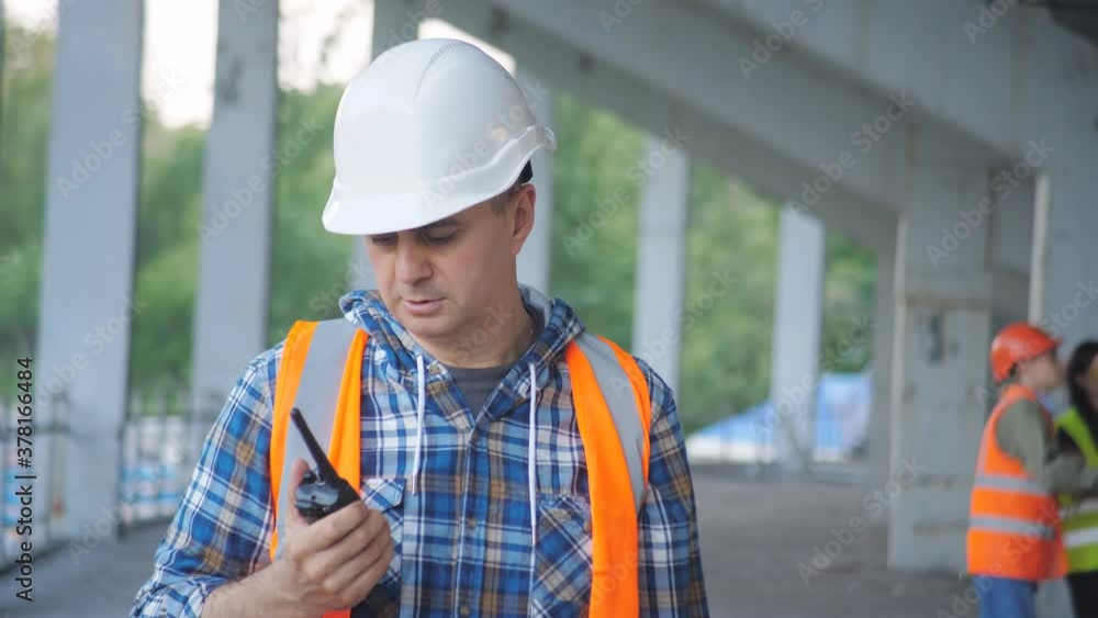 Close portrait of male builder in hardhat with walkie talkie or radio at construction site. Foreman wearing safety uniform and hardhat talking via walkie-talkie while standing at construction site Close portrait of male builder in hardhat with walkie talkie or radio at construction site. Foreman wearing safety uniform and hardhat talking via walkie-talkie while standing at construction site