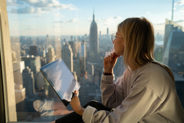 Thoughtful female digital nomad with modern touch pad looking in panoramic window while creation social content for sharing to web blog, millennial hipster girl with gadget for online communication
