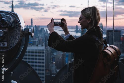 Millennial female traveller using cellphone camera for shooting video content visiting viewpoint observation with coin operated binocular for lookout on New York cityscape, touristic getaway