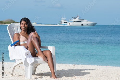 Brunette Woman applying sunscreen protection lotion sitting on white beach chair. Health concepts and skin care