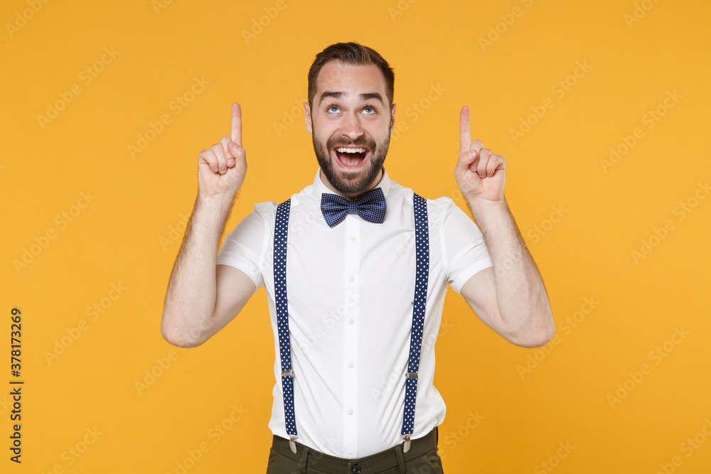 Fototapeta premium Cheerful excited young bearded man 20s in white shirt bow-tie suspender posing standing pointing index fingers up on mock up copy space isolated on bright yellow color wall background studio portrait.