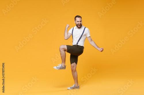 Full length side view portrait of joyful happy young bearded man 20s wearing white shirt suspender shorts posing clenching fists doing winner gesture isolated on bright yellow color background studio. © ViDi Studio