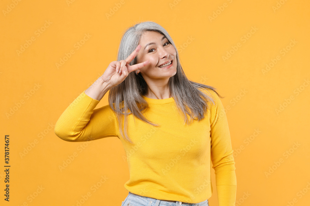 Smiling funny excited cheerful attractive gray-haired asian woman wearing casual clothes standing showing victory sign looking camera isolated on bright yellow colour background, studio portrait. Smiling funny excited cheerful attractive gray-haired asian woman wearing casual clothes standing showing victory sign looking camera isolated on bright yellow colour background, studio portrait.