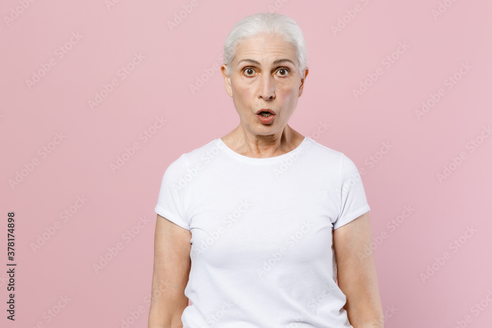 Shocked elderly gray-haired female woman 60s 70s wearing white blank empty casual t-shirt posing keeping mouth open looking camera isolated on pastel pink color wall background studio portrait. Shocked elderly gray-haired female woman 60s 70s wearing white blank empty casual t-shirt posing keeping mouth open looking camera isolated on pastel pink color wall background studio portrait.