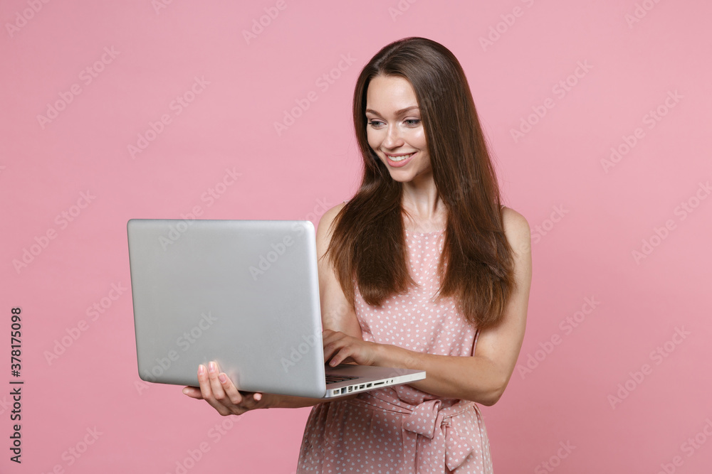 Smiling beautiful attractive young brunette woman 20s wearing pink summer dotted dress posing holding in hands working on laptop pc computer isolated on pastel pink color background studio portrait. Smiling beautiful attractive young brunette woman 20s wearing pink summer dotted dress posing holding in hands working on laptop pc computer isolated on pastel pink color background studio portrait.