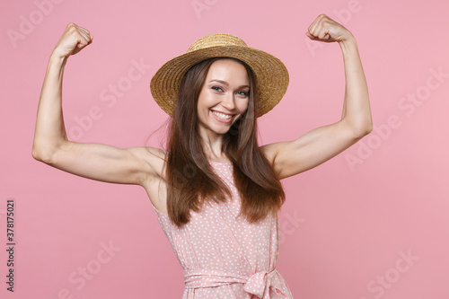 Smiling cheerful strong beautiful young brunette woman 20s wearing pink summer dotted dress hat posing showing biceps muscles looking camera isolated on pastel pink wall background studio portrait.