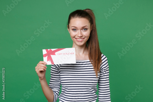 Smiling attractive beautiful young brunette woman 20s wearing striped casual clothes posing holding in hand gift certificate looking camera isolated on green color wall background studio portrait.