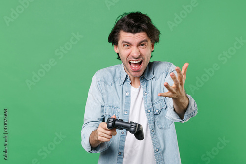Angry irritated screaming young brunet man 20s wearing casual clothes white t-shirt denim shirt posing standing play game with joystick spreading hands isolated on green background studio portrait.