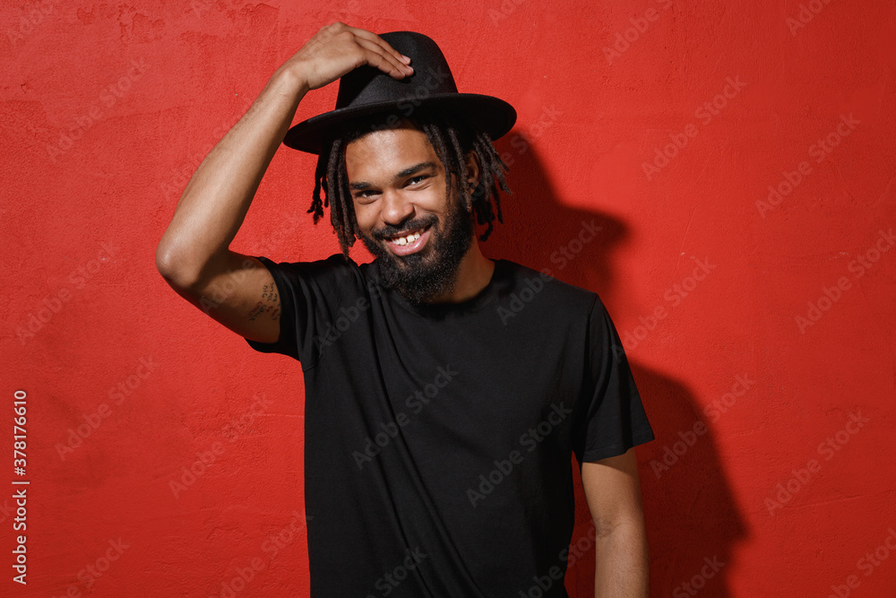 Smiling handsome attractive young african american man guy with dreadlocks 20s in black casual t-shirt posing hold hat on head looking camera isolated on bright red color background studio portrait. Smiling handsome attractive young african american man guy with dreadlocks 20s in black casual t-shirt posing hold hat on head looking camera isolated on bright red color background studio portrait.