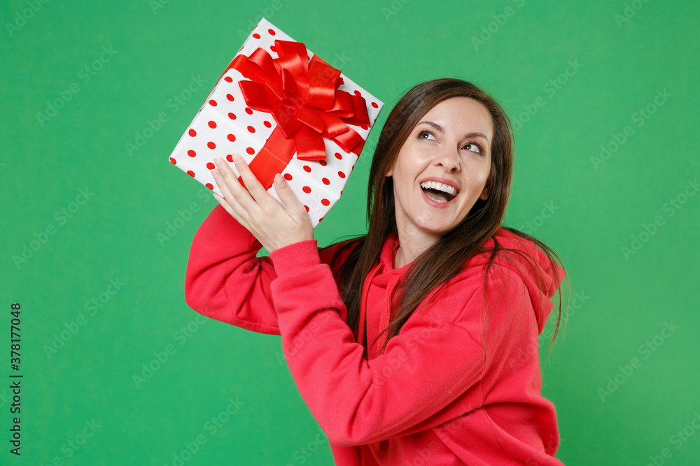 Cheerful curious funny young brunette woman in red casual streetwear hoodie posing celebrating hold red present box with gift ribbon bow looking up isolated on green color background studio portrait. Cheerful curious funny young brunette woman in red casual streetwear hoodie posing celebrating hold red present box with gift ribbon bow looking up isolated on green color background studio portrait.