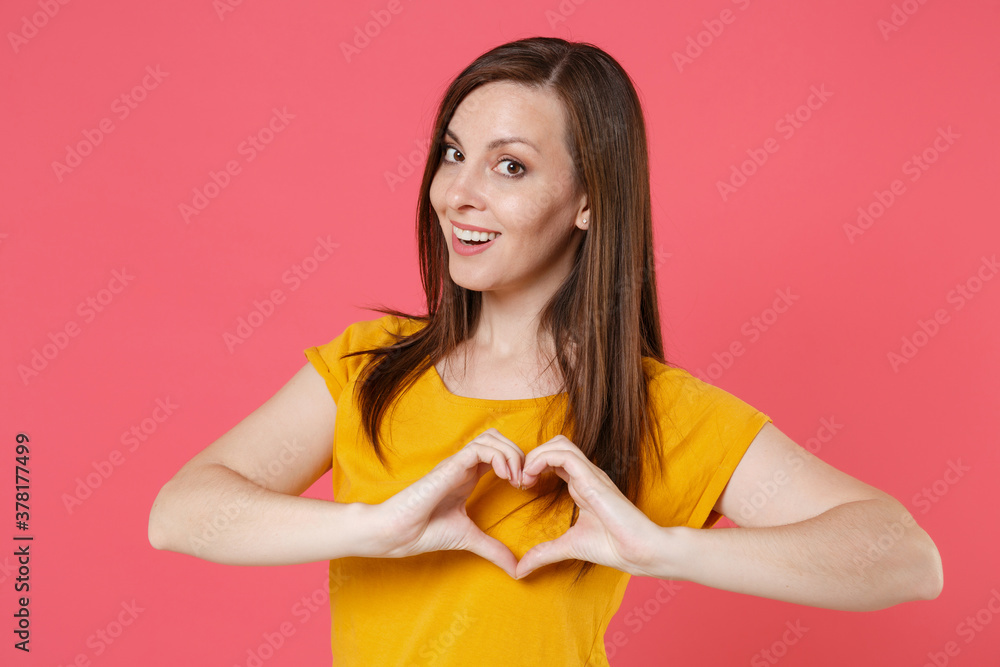 Smiling pretty young brunette woman 20s wearing yellow casual t-shirt posing standing showing shape heart with hands, heart-shape sign looking camera isolated on pink color background studio portrait. Smiling pretty young brunette woman 20s wearing yellow casual t-shirt posing standing showing shape heart with hands, heart-shape sign looking camera isolated on pink color background studio portrait.